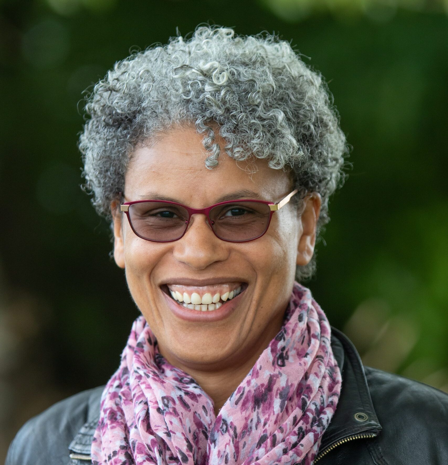 Claire Bankole Headshot: A smiling person with curly hair and glasses, wearing a jacket and a pink patterned scarf.